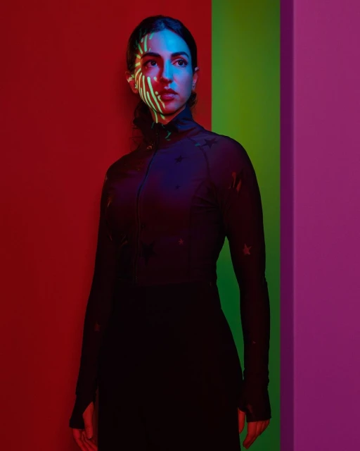 Portrait of a woman in a black turtleneck and skirt stands against a vibrant multicolor wall with a striped light pattern on her face.